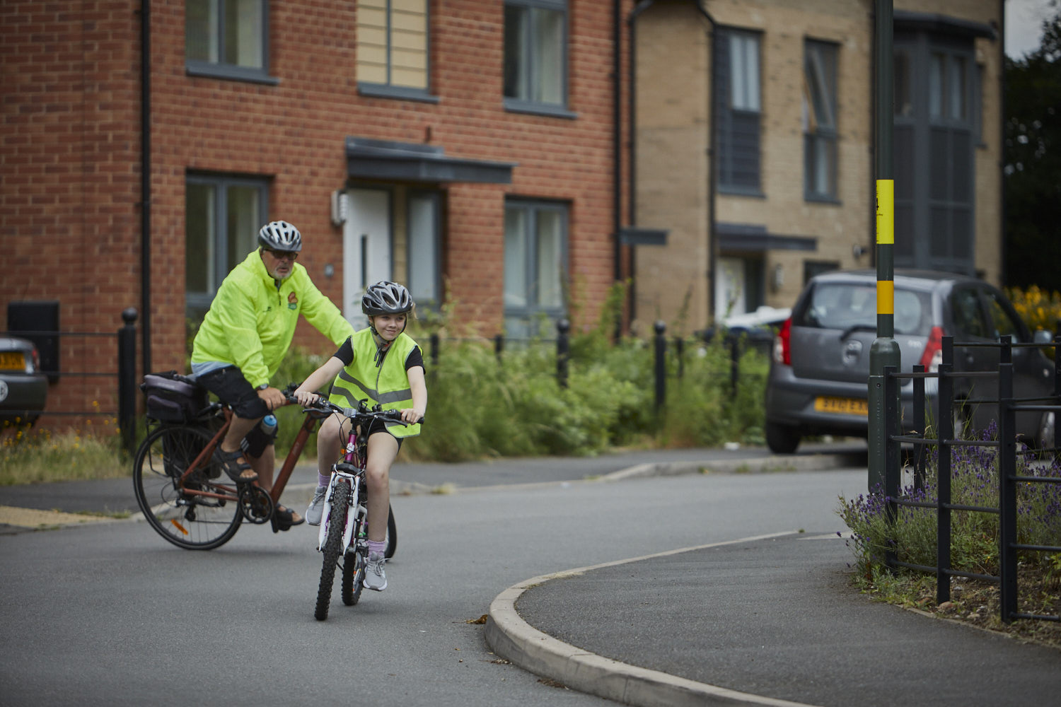 Smiling child in high visibility clothing and helmet rides bicycle on the road with adult watching