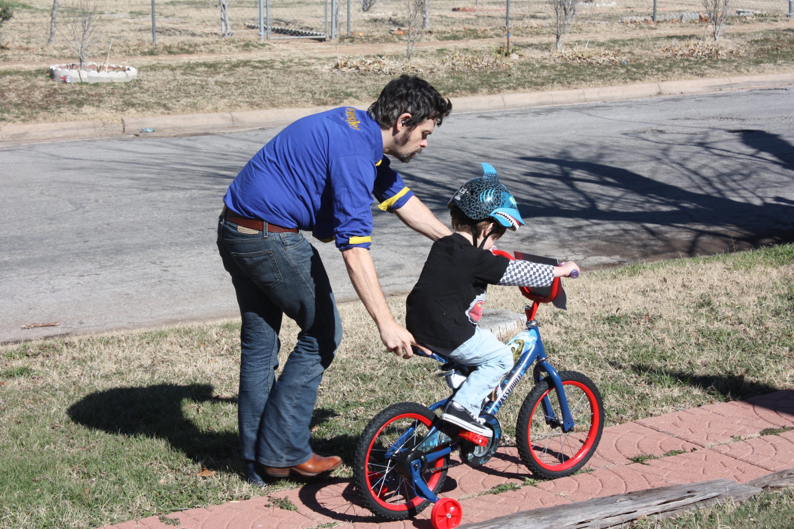 Father teaching son to ride and pushing his bicycle down pathway near roadside