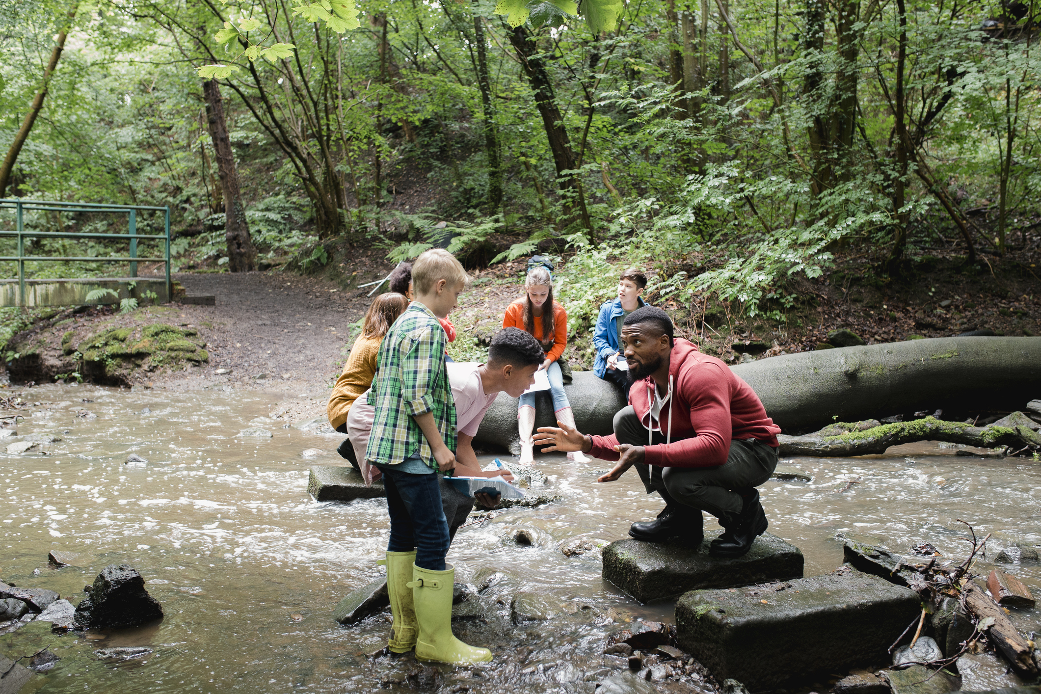 Crouching man teaches group of inquisitive children next to river in wooded forest