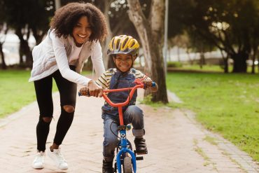 Smiling mother helps young son ride bicycle down pathway during daytime