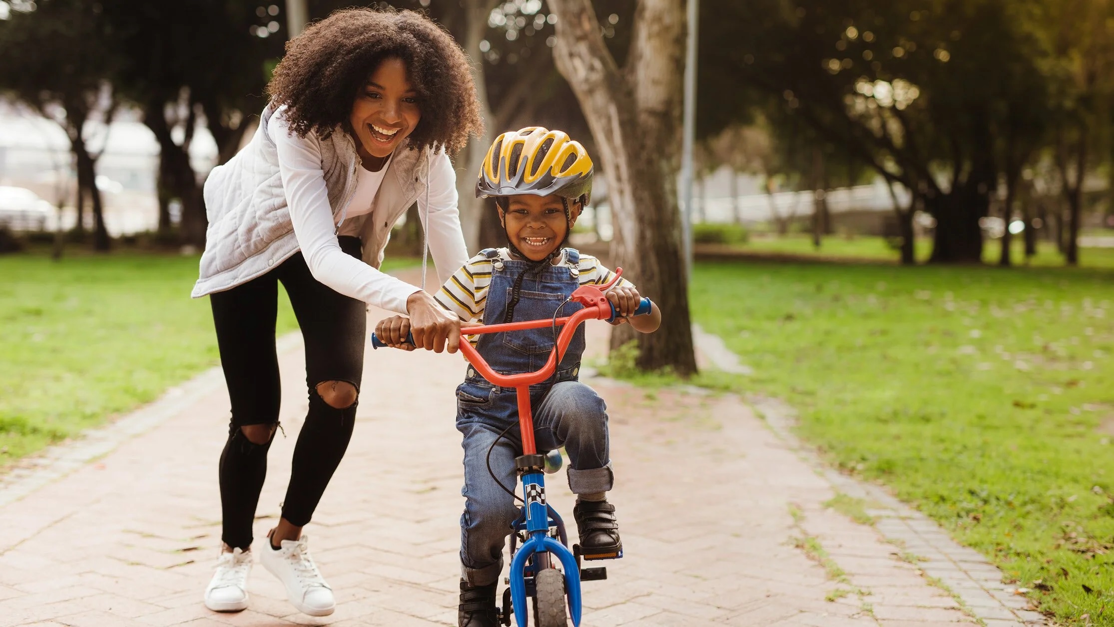 Smiling mother helps young son ride bicycle down pathway during daytime